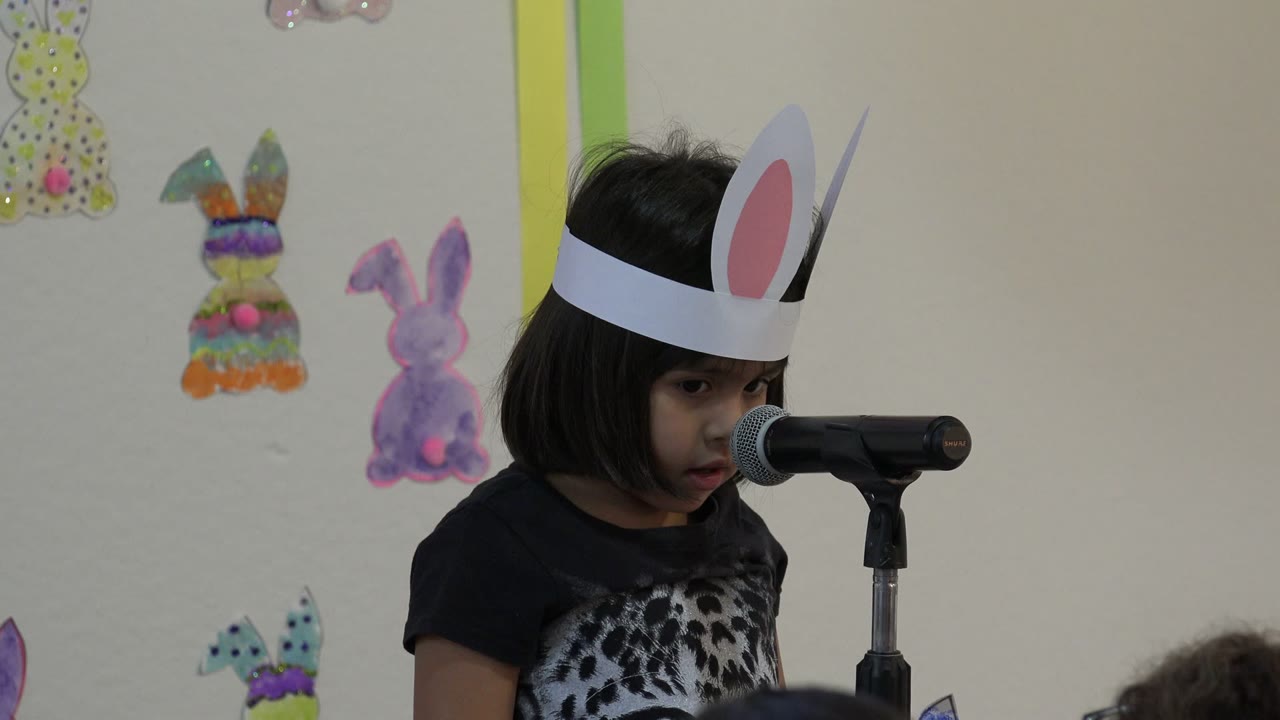 Young girl wearing paper bunny ears speaks into a microphone.
