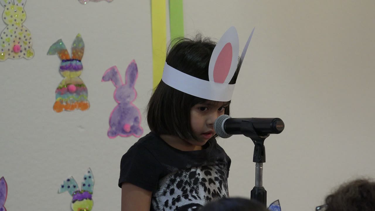 At 0:01, the child remains near the microphone, her face partly turned downward, with colorful rabbit cutouts visible on the wall in Cabo San Lucas, Mexico.