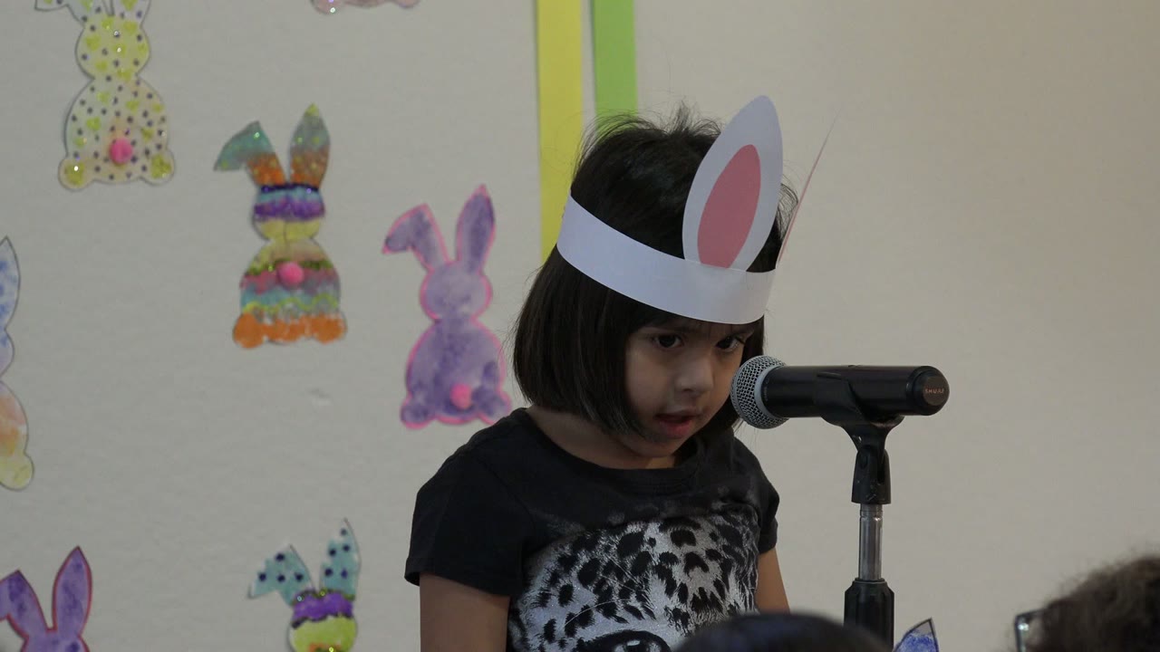 At 0:01, the child looks downward beside the microphone, wearing white bunny ears in front of handmade Easter bunny decorations in Cabo San Lucas, Mexico.