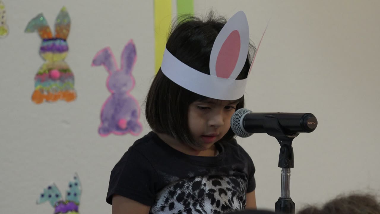 A young girl in Cabo San Lucas, Mexico wears white paper bunny ears and looks down beside a black microphone, with colorful bunny decorations on the wall behind her.