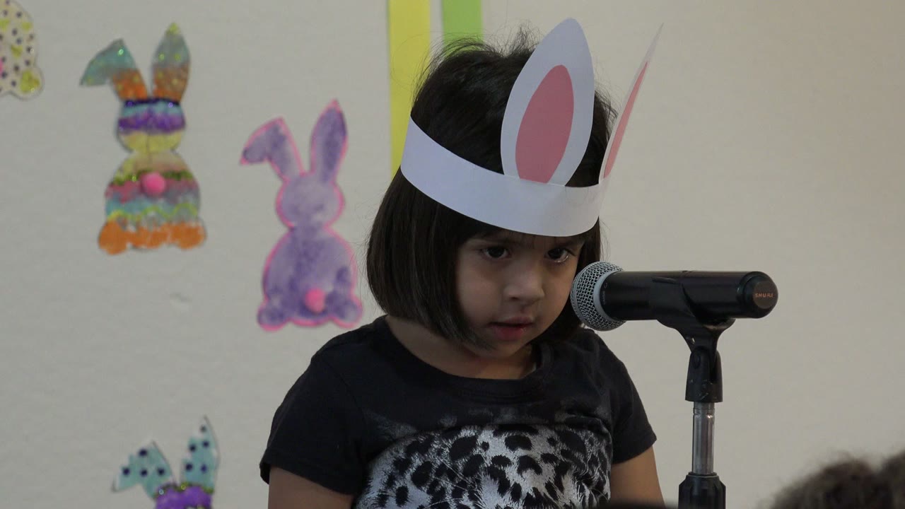 The girl in Cabo San Lucas, Mexico faces forward near the microphone, her white bunny-ear headband prominent against the pale decorated wall.