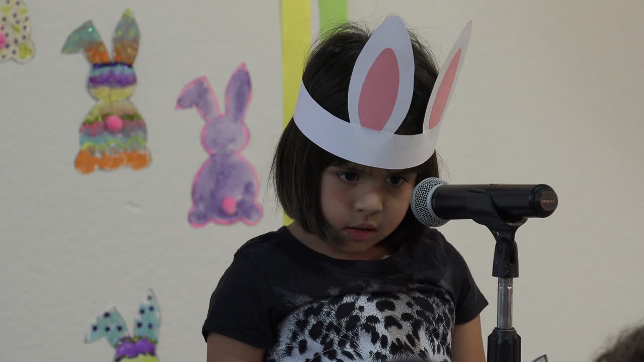 Little girl wearing paper bunny ears stands beside a microphone.