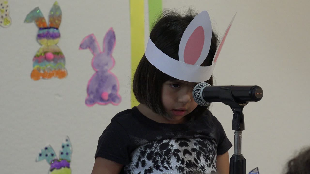 The child in Cabo San Lucas, Mexico looks down while standing beside the microphone, with colorful rabbit drawings visible on the wall behind her.