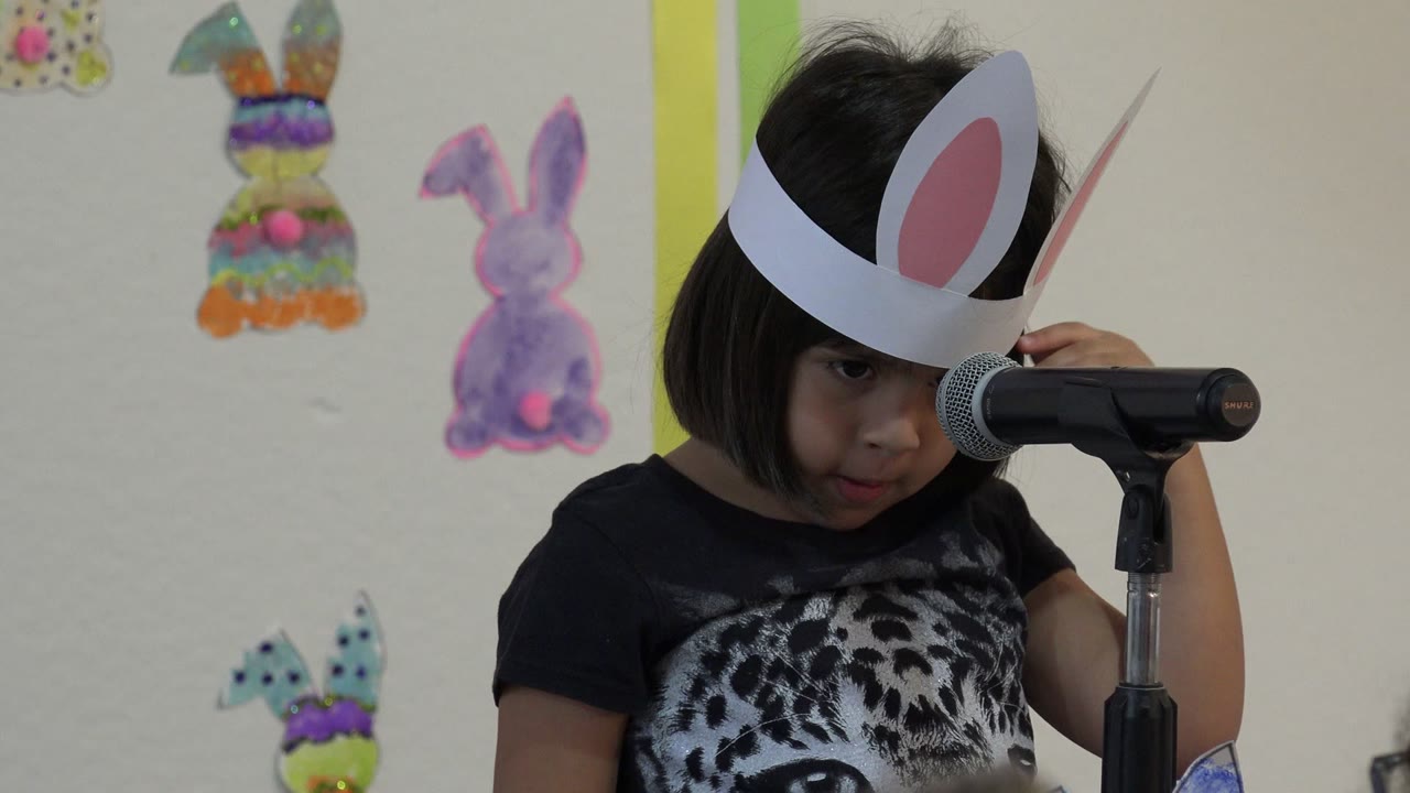 A young girl in Cabo San Lucas, Mexico wears paper bunny ears and stands close to a black microphone with rabbit drawings on the classroom wall behind her.