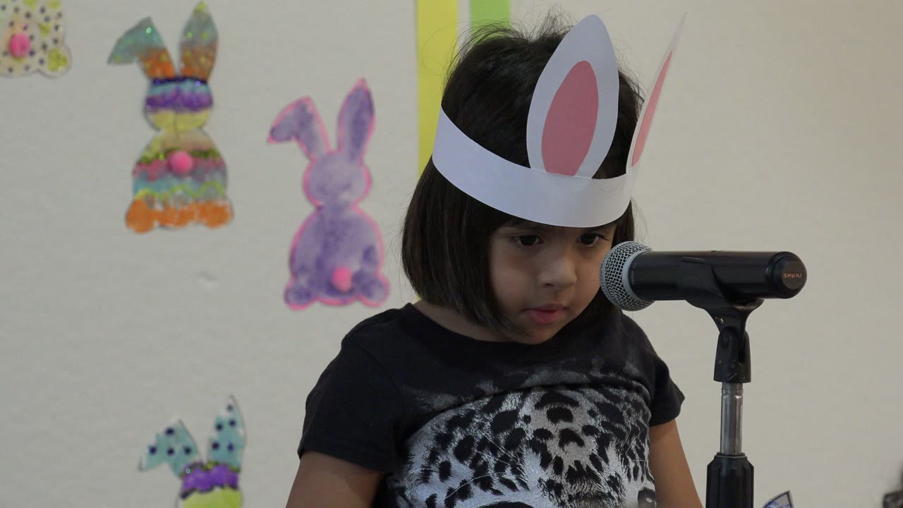 The child looks downward beside the microphone, with the white-and-pink bunny ears high in frame and painted rabbit decorations behind her.