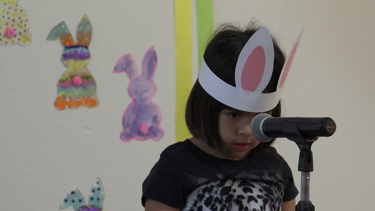 A young girl wearing paper bunny ears stands beside a black microphone during an Easter school presentation in Cabo San Lucas, Mexico, with handmade bunny decorations on the wall behind her.