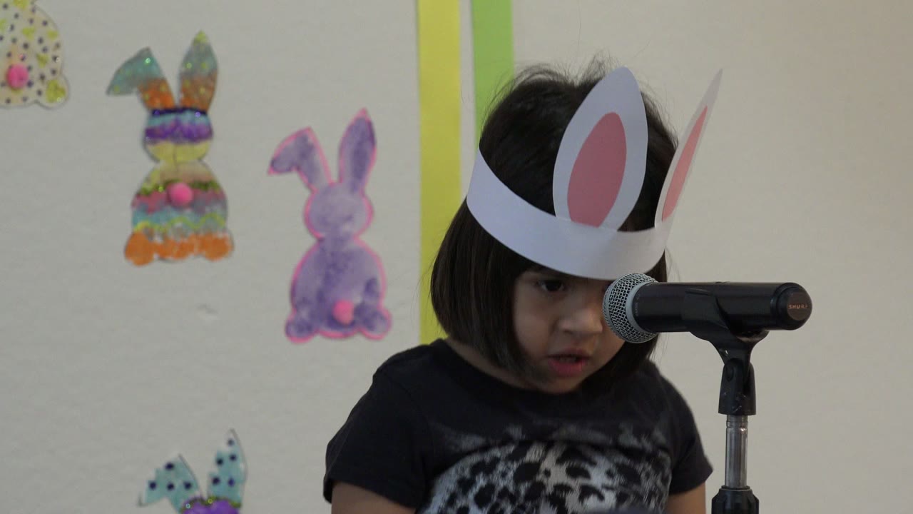 The child in white paper bunny ears looks downward near the microphone, with colorful bunny cutouts and green vertical trim visible on the school wall in Cabo San Lucas, Mexico.
