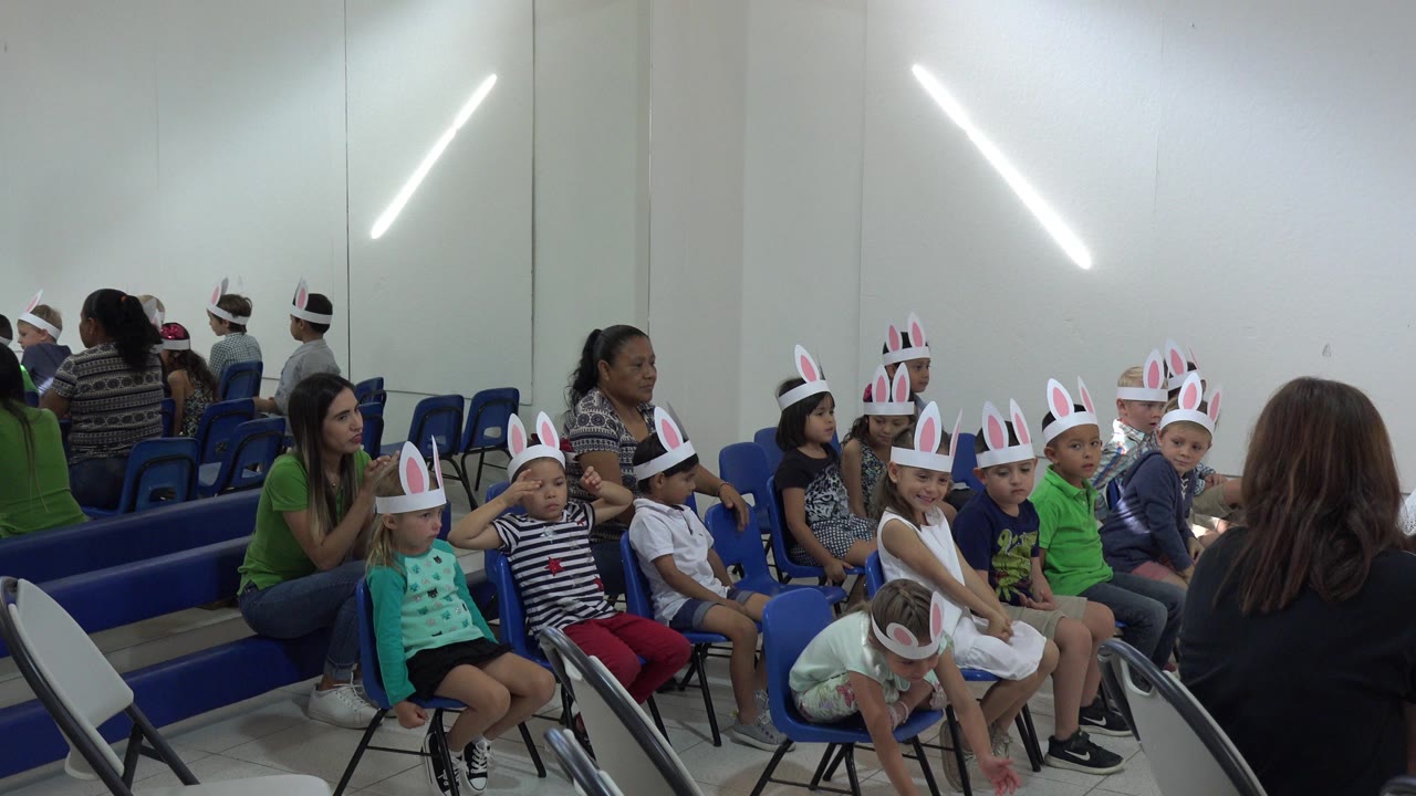 Children in rabbit ears sit in blue chairs with adults nearby.