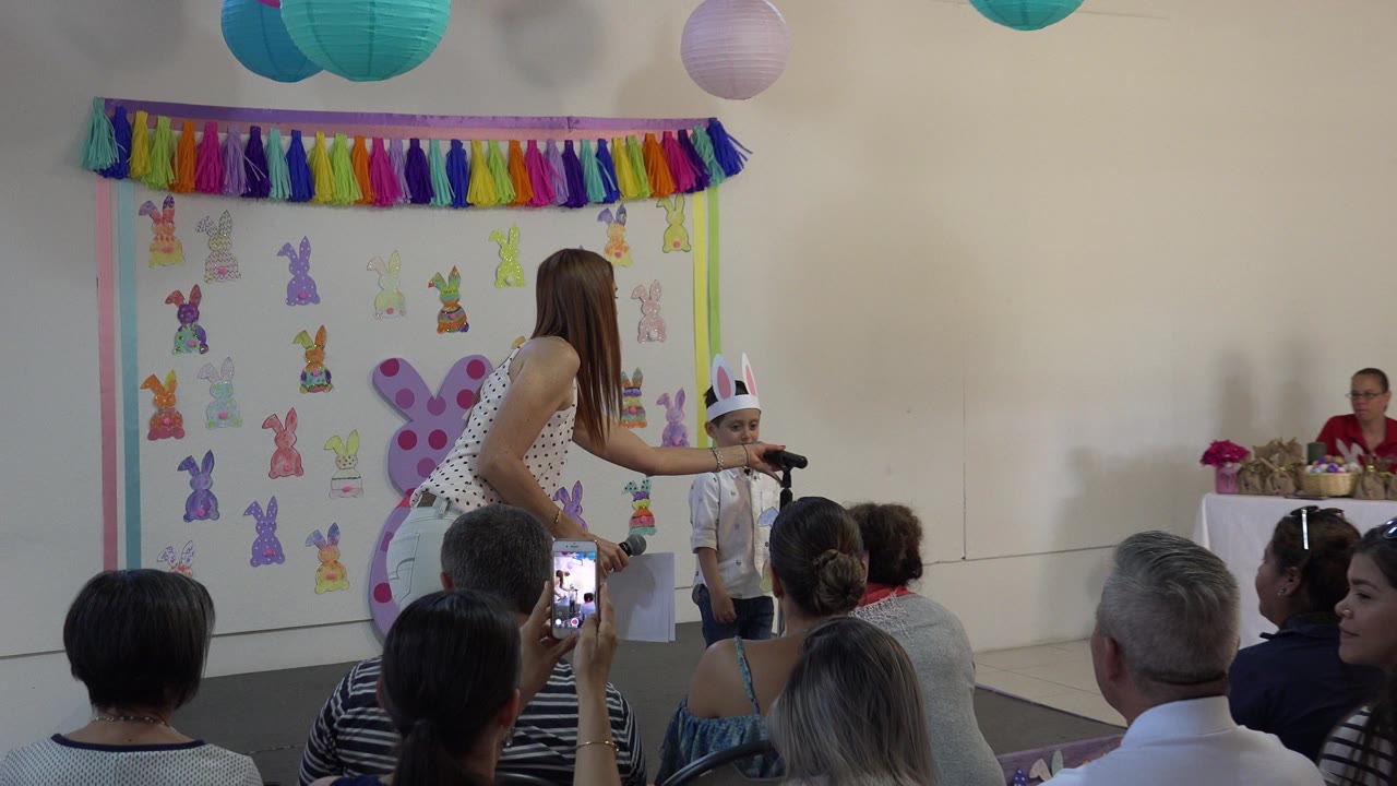 Boy in rabbit ears at a classroom microphone as an adult adjusts the setup.