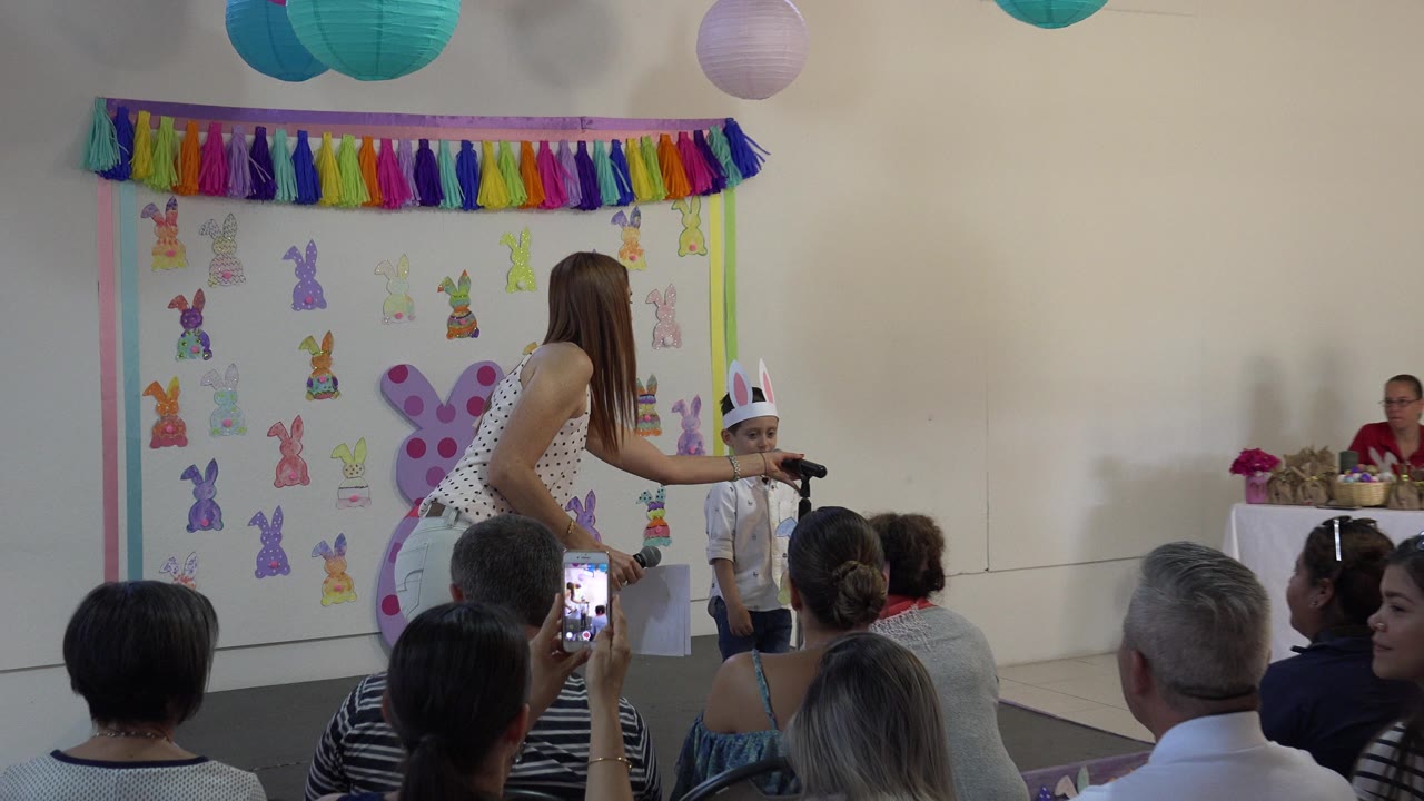 In Cabo San Lucas, Mexico, the boy waits at the microphone in front of a bunny-decorated wall while an adult stands close and audience heads fill the lower frame.