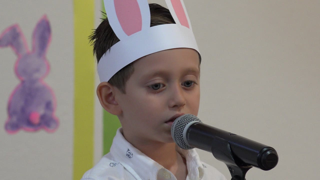 The boy in bunny ears looks down toward the microphone in a close-up from the Cabo San Lucas school concert, with a painted bunny decoration on the wall.
