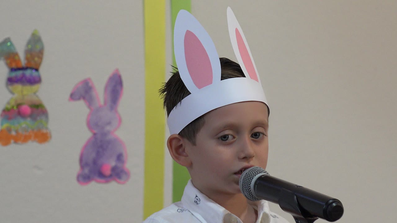 At 0:00, a boy in white paper bunny ears faces a black microphone during an Easter school performance in Cabo San Lucas, Mexico, with bunny decorations behind him.