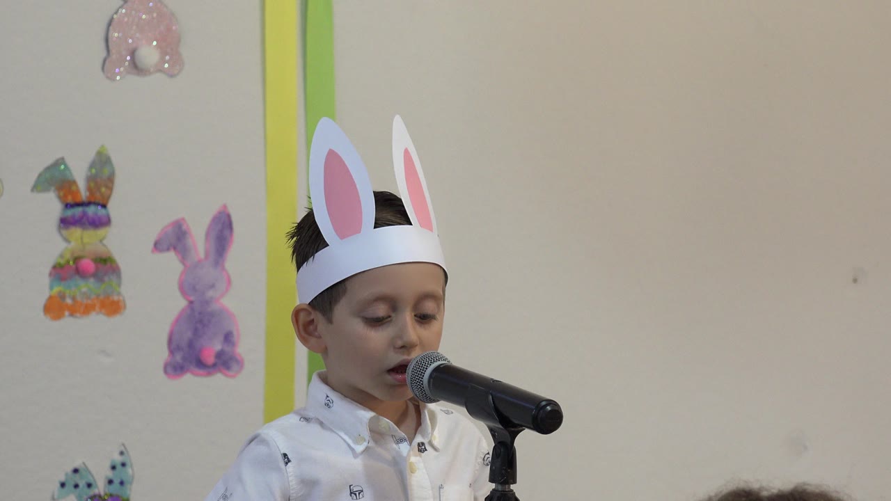At 0:00, a young boy in paper bunny ears stands at a black microphone during a Cabo San Lucas school event, with handmade rabbit decorations on the wall behind him.