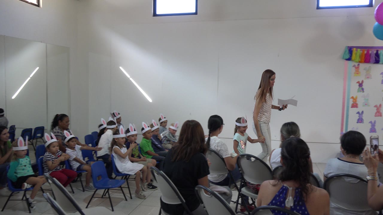 Children in rabbit-ear headbands sit before a decorated presentation wall.