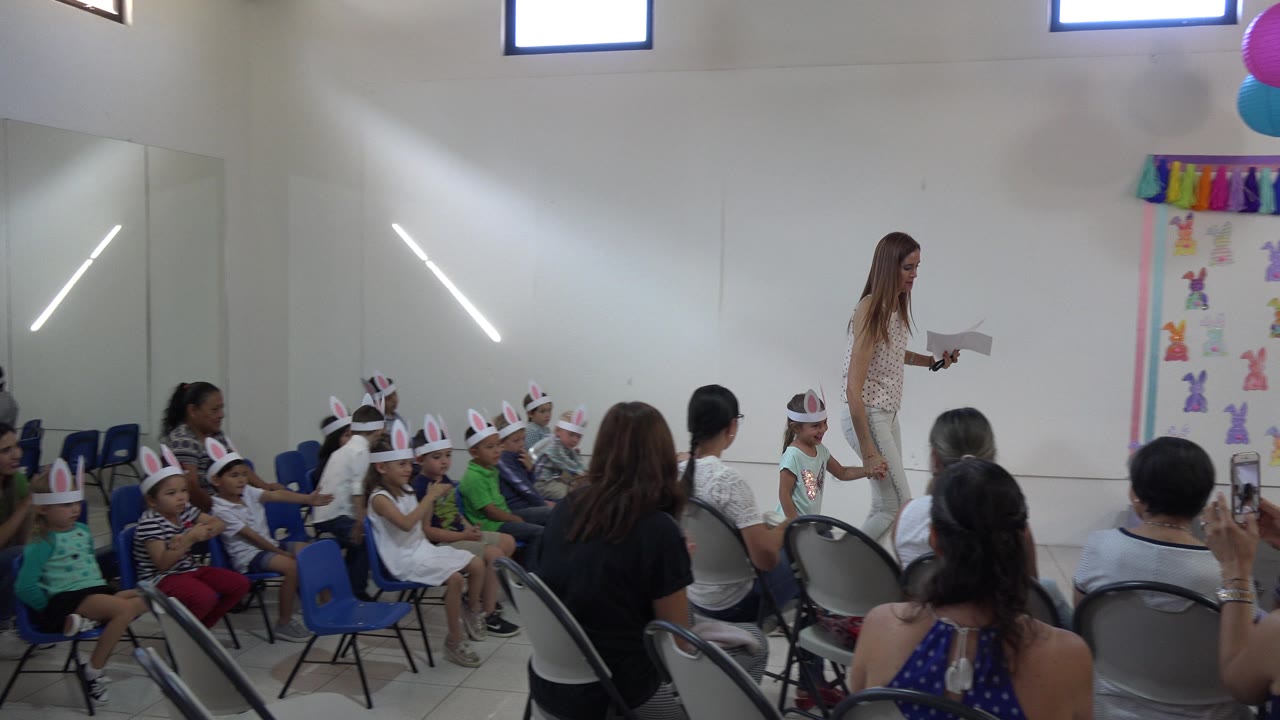 A woman stands at the front of an indoor Cabo San Lucas children's event while children in rabbit ears and seated adults face the decorated presentation area.
