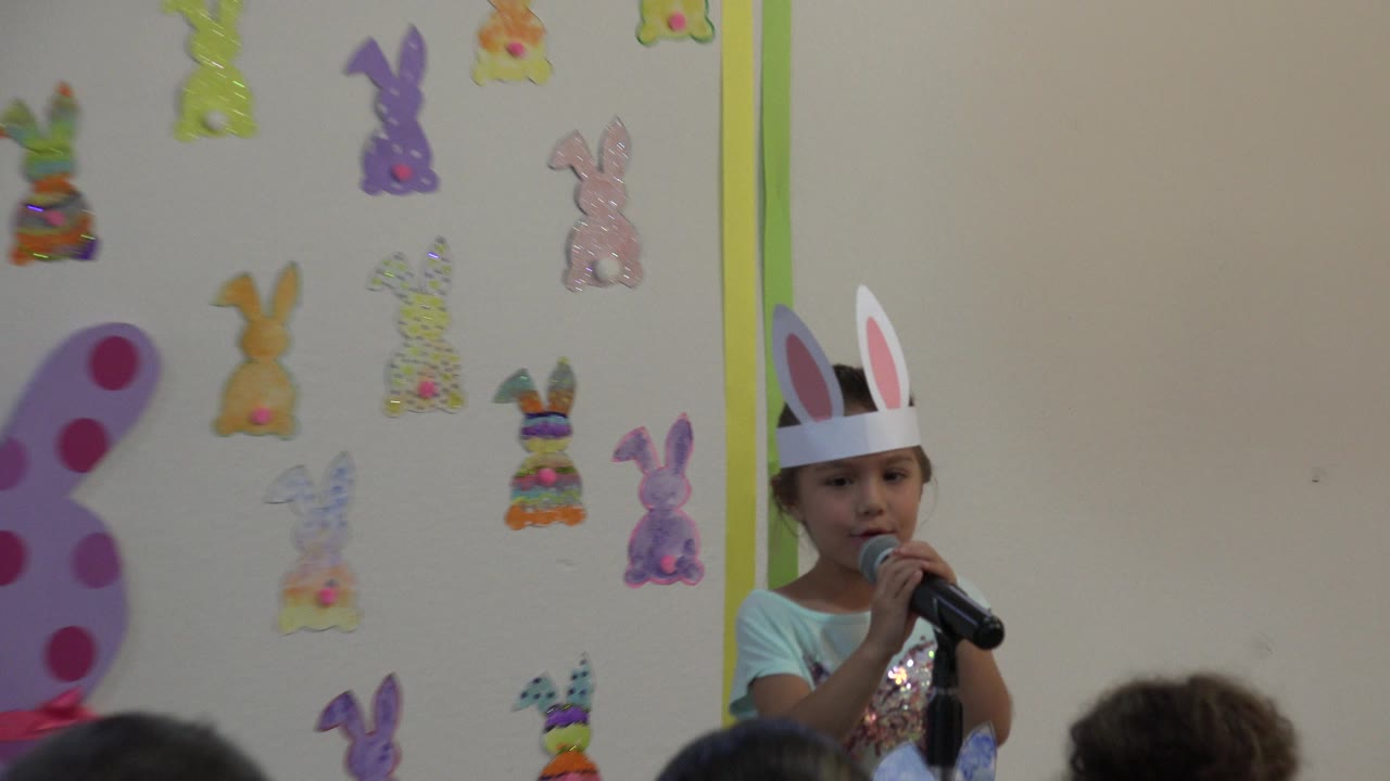 Wide Cabo San Lucas 2019 frame of a young girl in white rabbit ears speaking into a black microphone beside colorful rabbit decorations on an indoor wall.