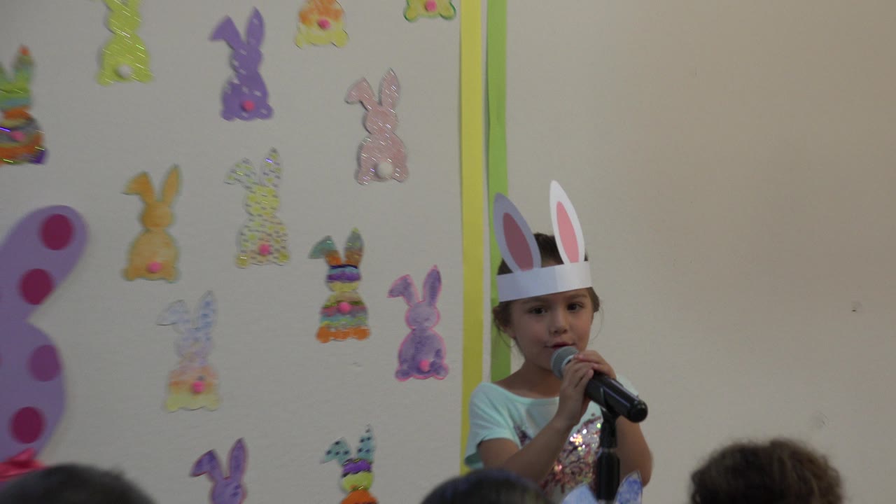 Cabo San Lucas 2019 frame with the girl centered lower in the shot, holding a black microphone while pastel rabbit cutouts fill the wall behind her.