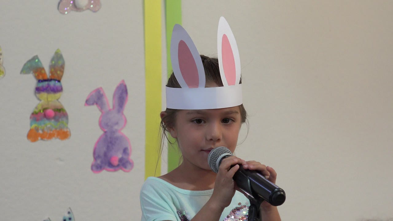 Young girl in bunny ears holding a microphone near Easter bunny decorations.