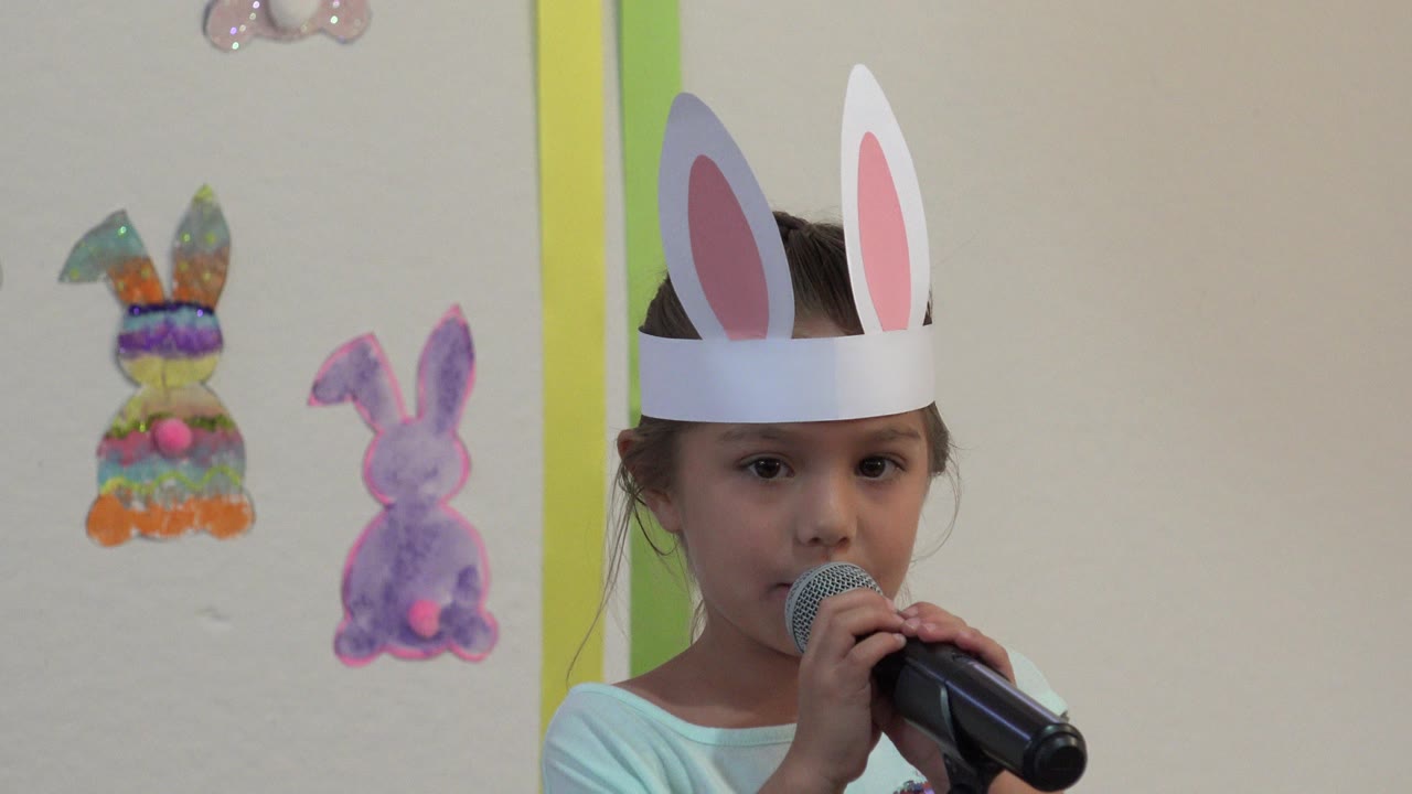 At 0:00, a young girl in Cabo San Lucas wears paper bunny ears and holds a microphone near her mouth, with colorful paper bunny decorations on the wall behind her.