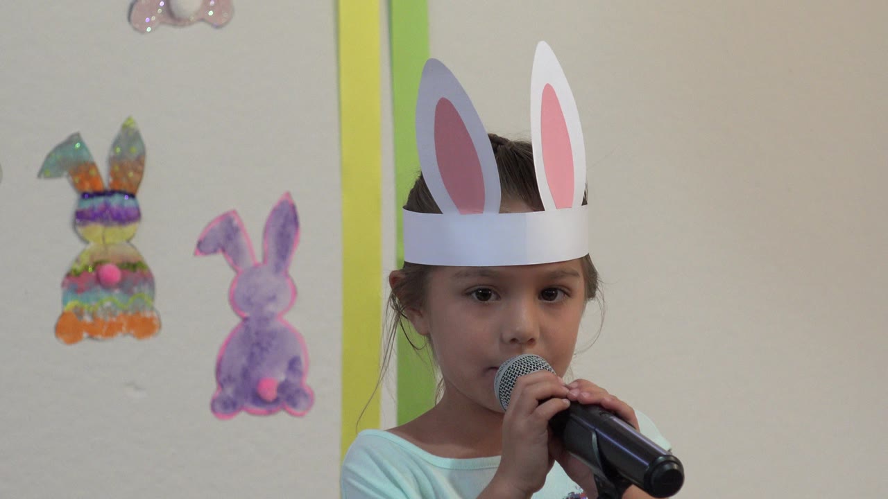 At 0:01, the girl remains centered in close framing, looking forward while singing into the handheld microphone against the decorated indoor wall in Cabo San Lucas.