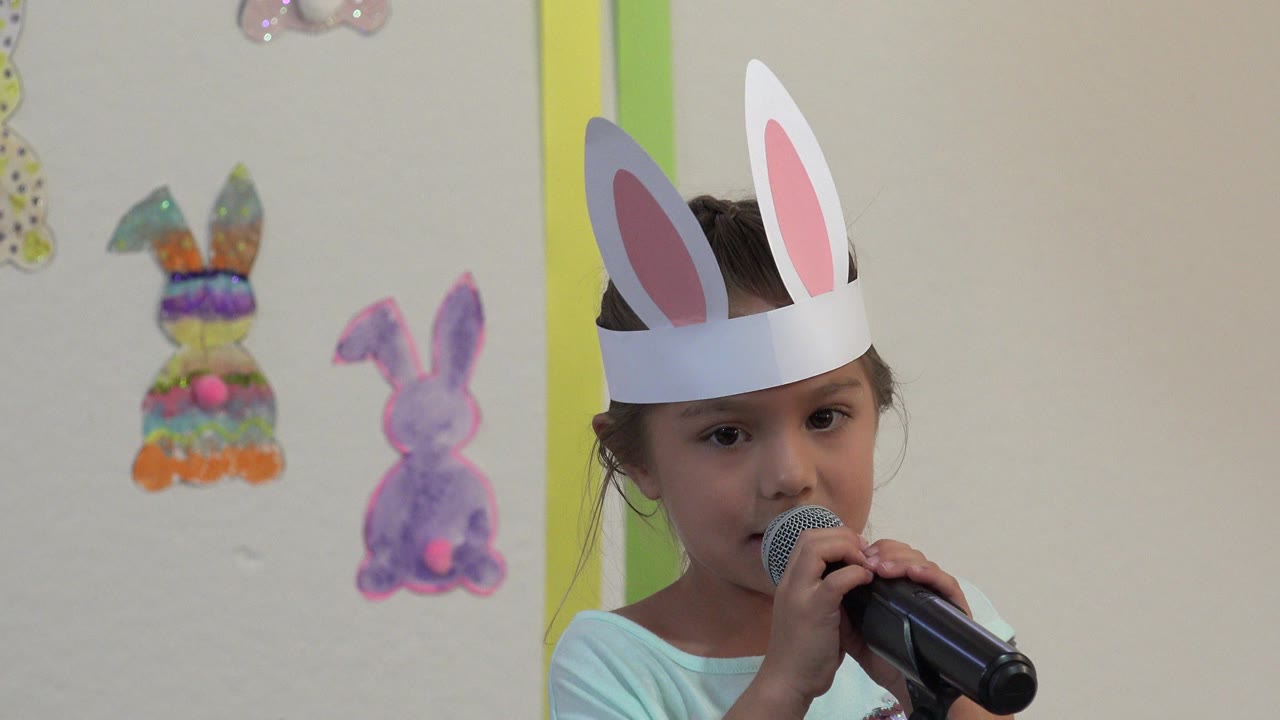 A child in paper bunny ears speaks into a handheld microphone during an Easter school presentation in Cabo San Lucas, Mexico, with bunny decorations behind them.