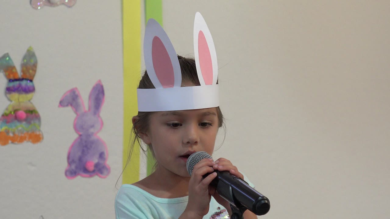 Young girl in paper bunny ears holding a microphone indoors.