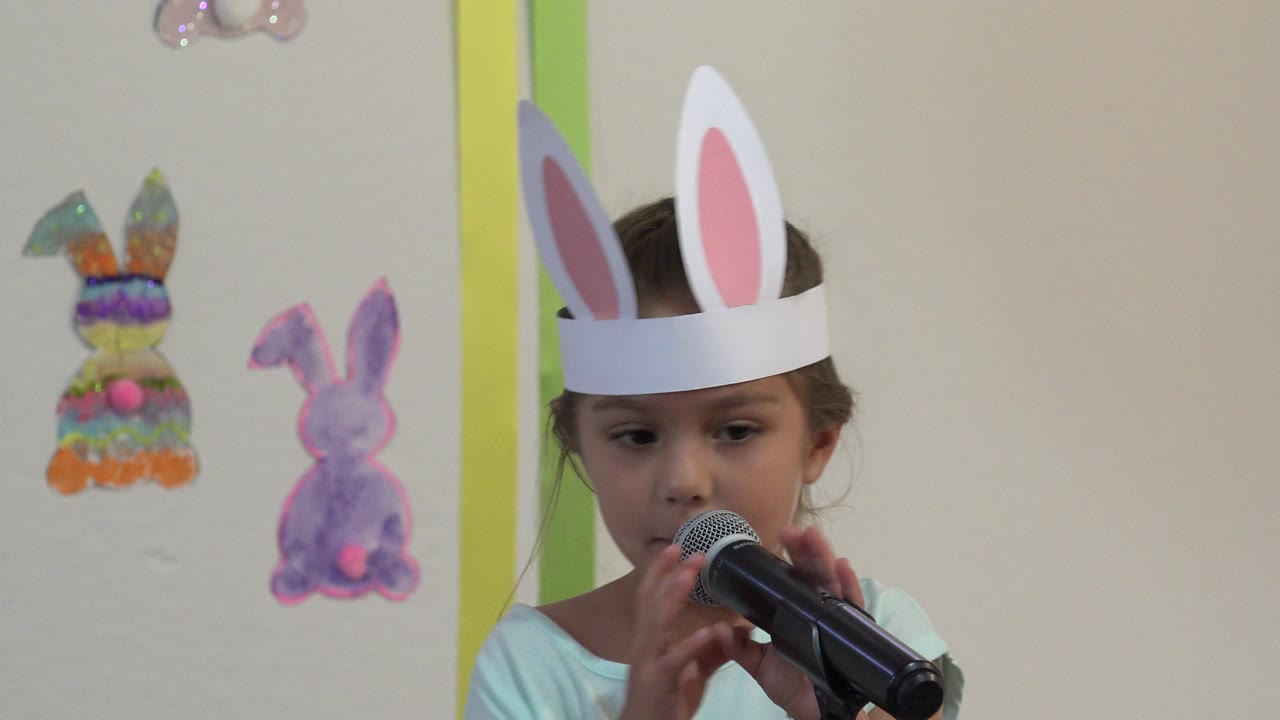 Child in bunny ears speaking into a microphone in a decorated classroom.