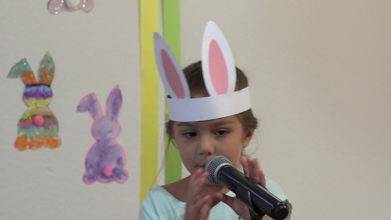 The child presenter remains centered at the microphone in the Cabo San Lucas classroom, with hands near the mic and colorful bunny cutouts visible on the wall.