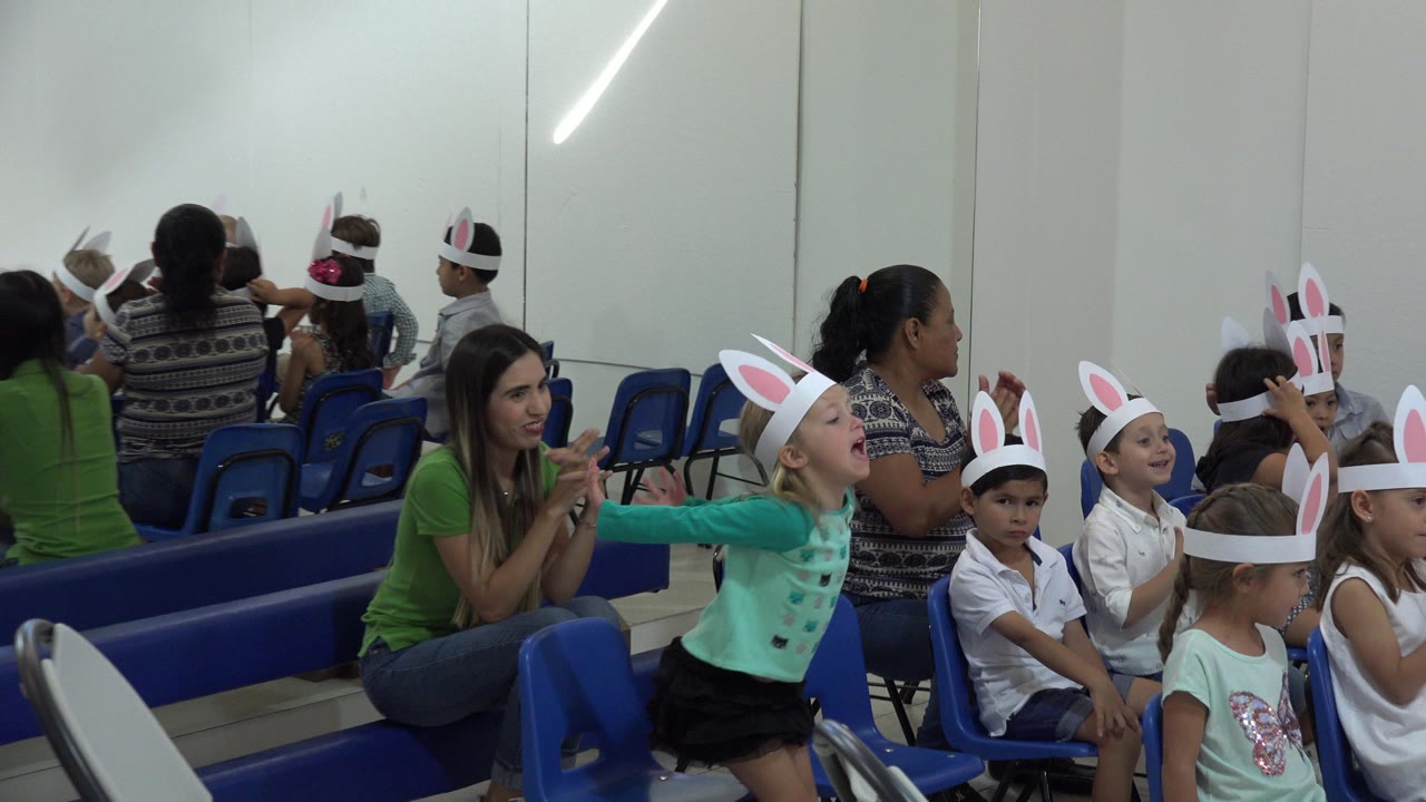 Children in paper bunny ears sit on blue chairs beside adults in an indoor room.