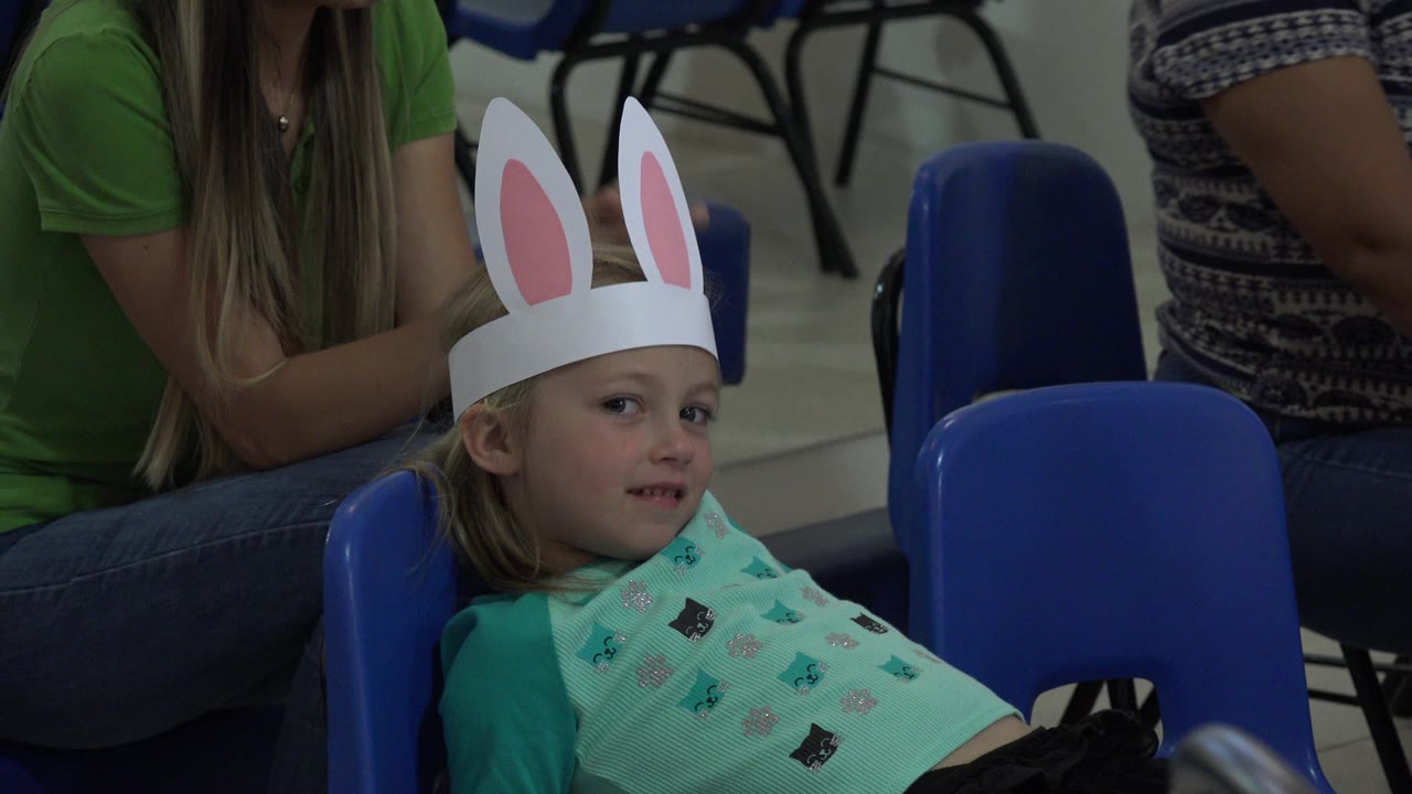 At 0:01, the girl remains reclined in the blue chair, facing the camera with her white rabbit-ear headband prominent and rows of chairs behind her.