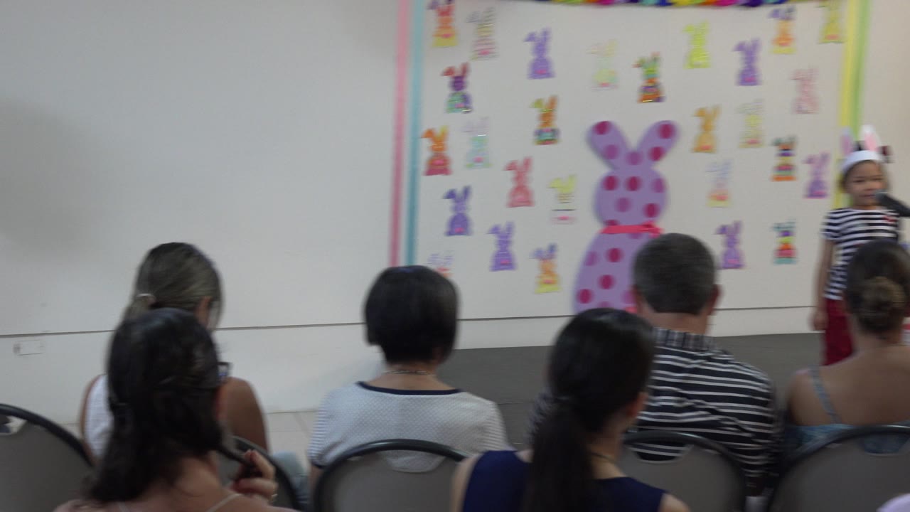 Child wearing bunny ears stands at a microphone before seated adults.