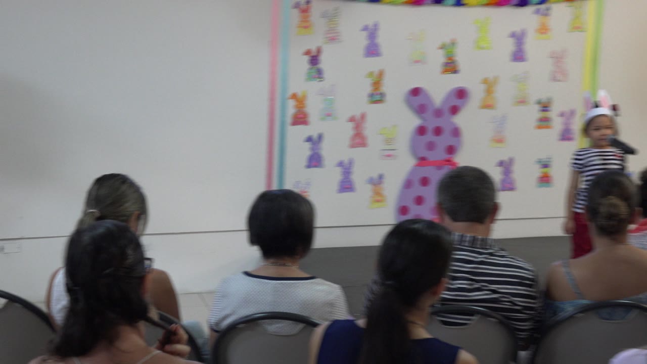 A young child at a microphone is centered beyond the seated audience, with a large pink bunny decoration and smaller drawings on the wall behind the performance area in Cabo San Lucas, Mexico.