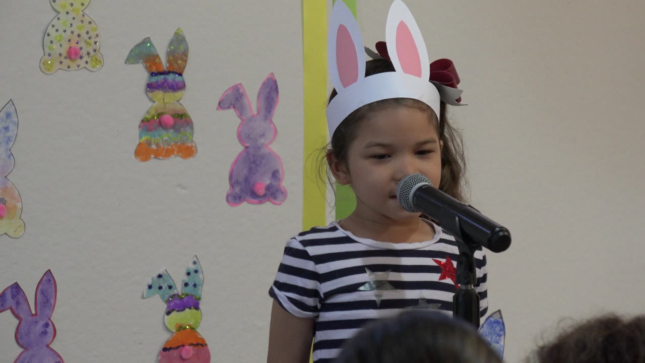 Young girl in bunny ears singing into a microphone in a classroom.