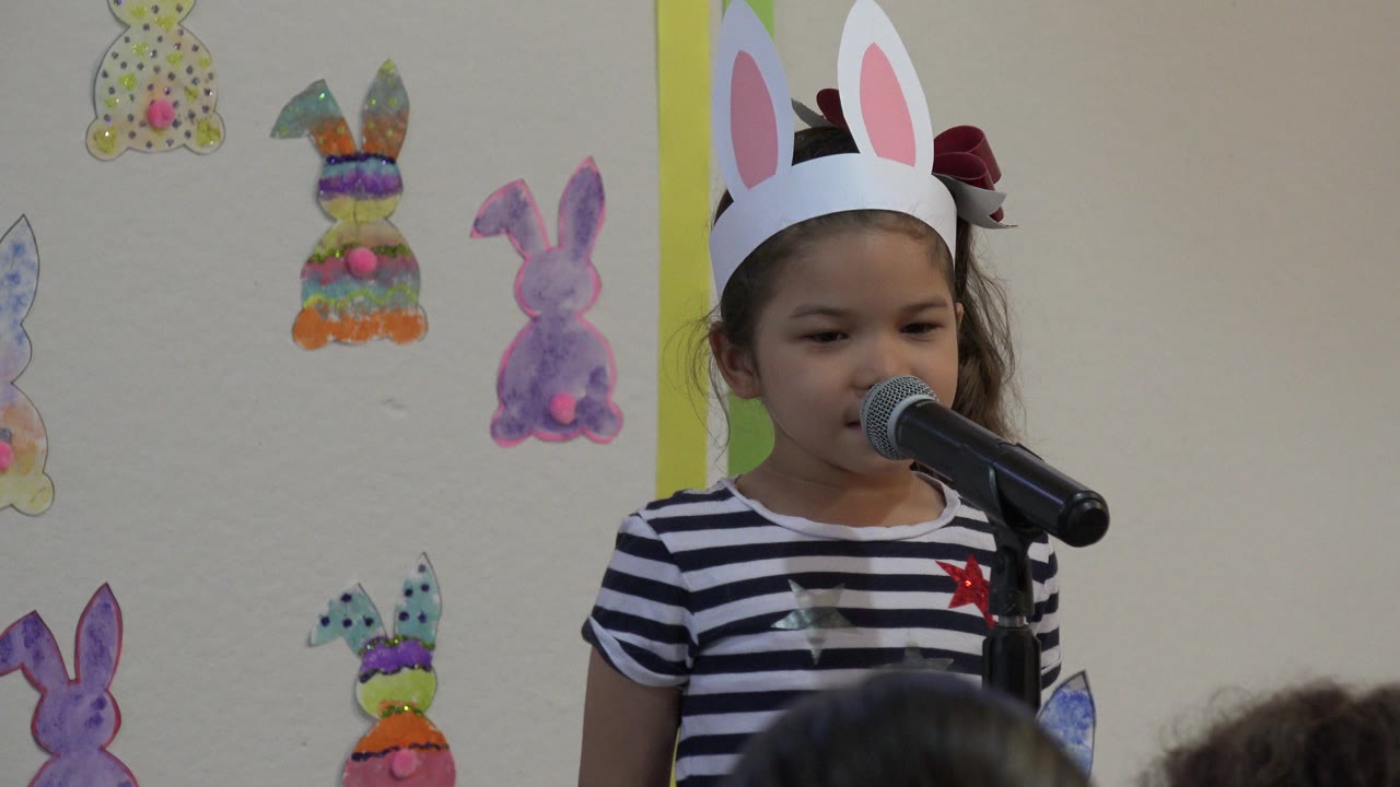 At 0:01, the child remains behind the microphone, framed slightly left of center with paper bunny decorations visible on the classroom wall in Cabo San Lucas.