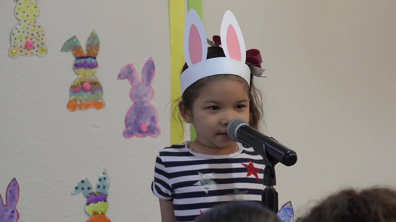 At 0:01, the girl faces slightly toward the microphone, her bunny ears high in frame, with colorful Easter decorations behind her and a few audience heads at the bottom.