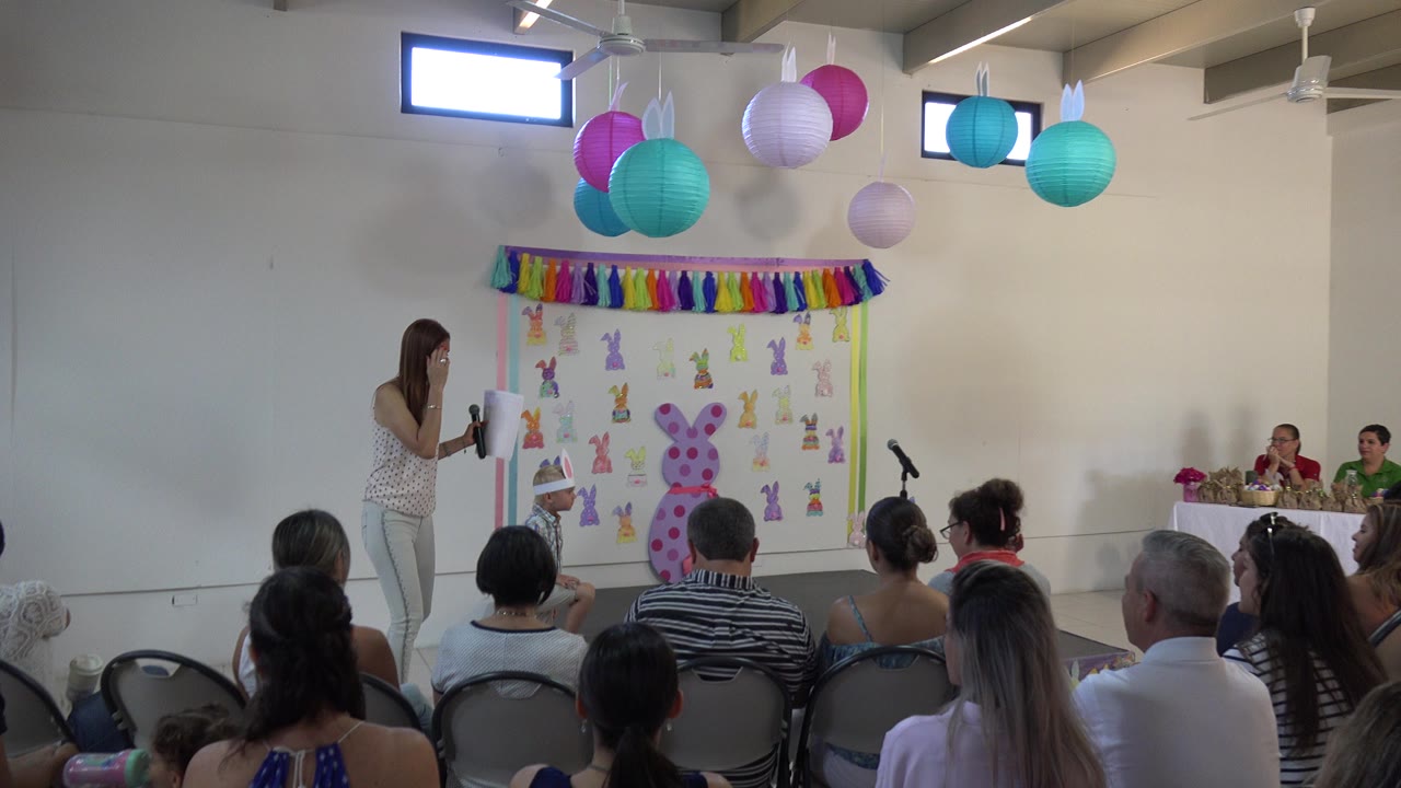 Teacher addresses seated audience in a decorated school hall.