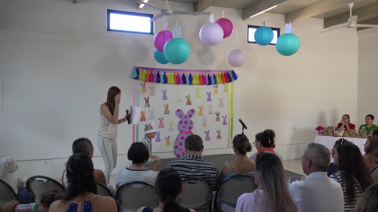 The teacher remains near the front of the Cabo San Lucas hall, holding papers beside a microphone with pastel lanterns overhead and bunny-themed decorations behind her.