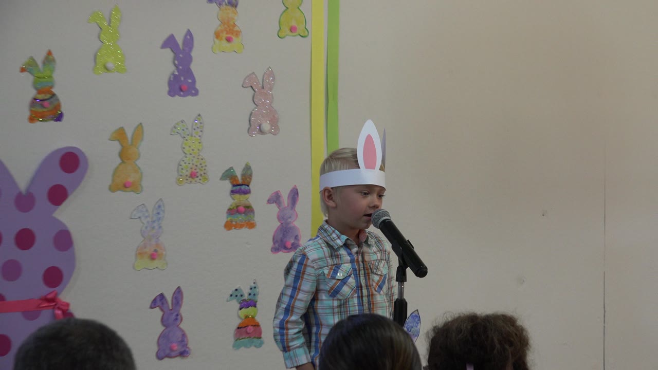 Young boy in paper bunny ears speaking at a microphone before an audience.