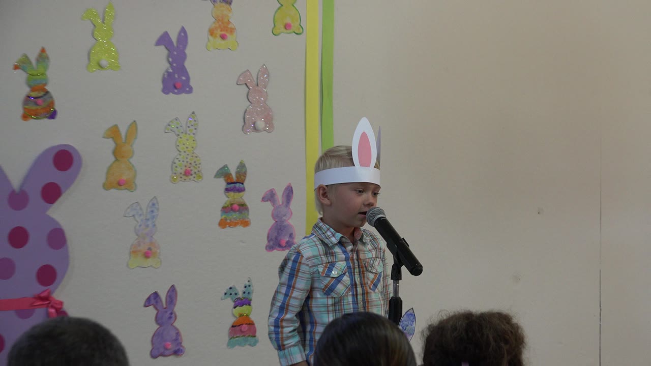 The boy in handmade bunny ears speaks into the microphone in Cabo San Lucas, Mexico, framed wider with audience heads at the bottom and paper bunny decorations behind him.