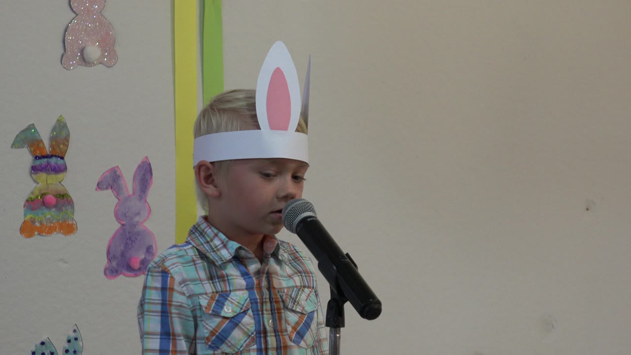 A young boy in Cabo San Lucas, Mexico wears white bunny ears and stands beside a black microphone with paper rabbit decorations on the wall behind him.