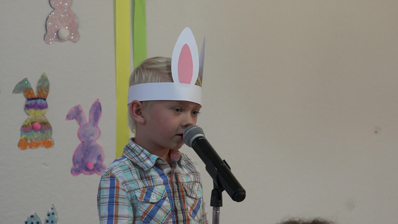 Boy in bunny ears speaking with colorful rabbit decorations behind him.