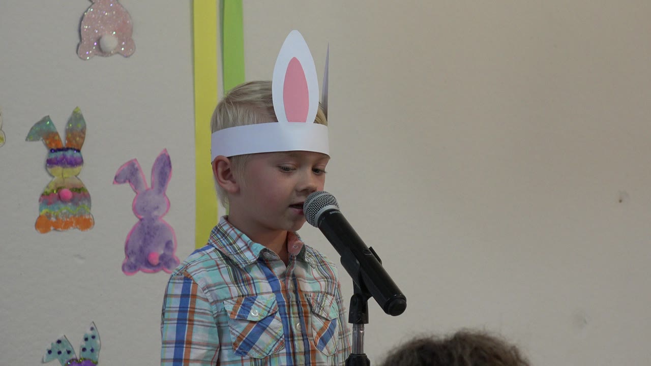 Boy in rabbit ears speaking at a microphone indoors.