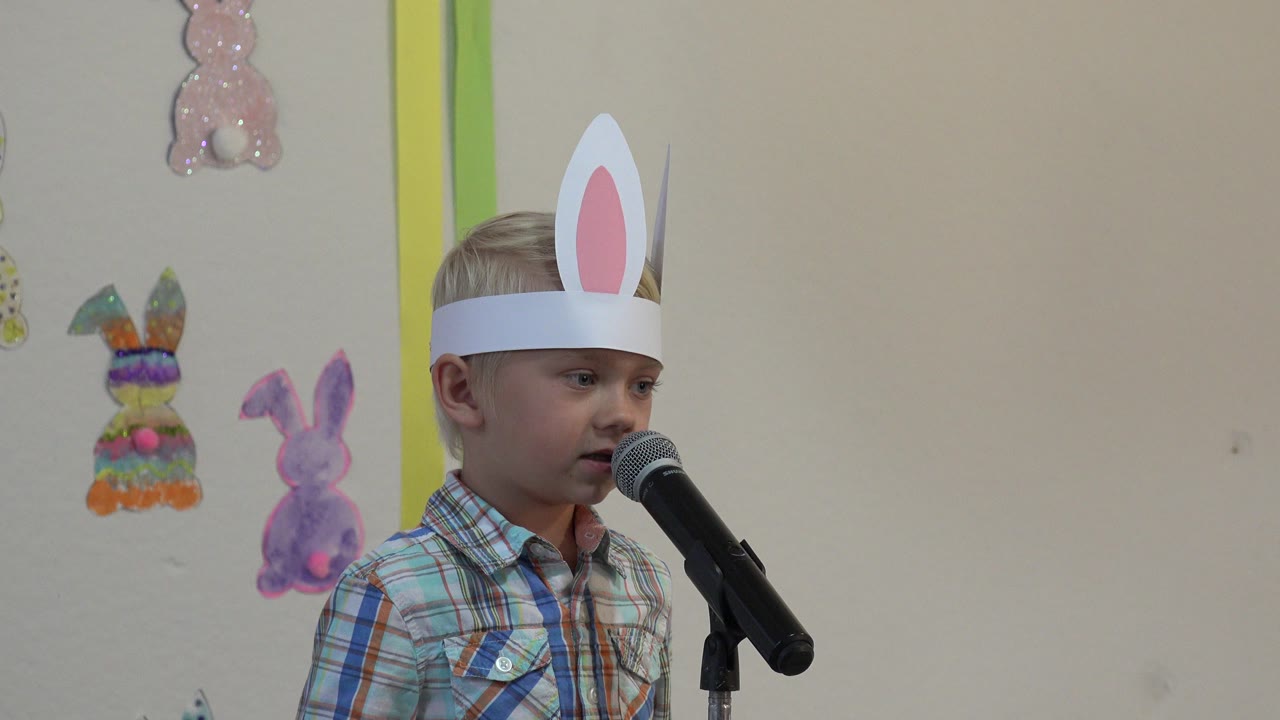 Boy wearing paper bunny ears speaks into a microphone near Easter decorations.
