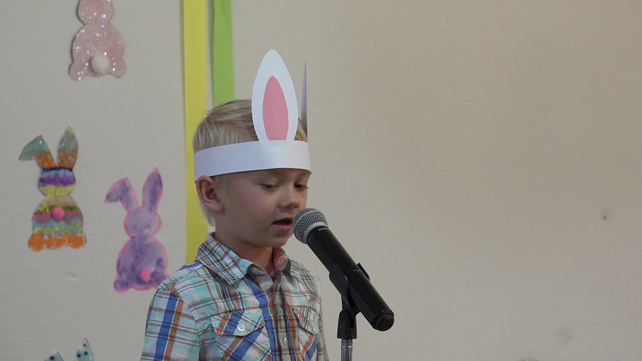 Young boy in paper bunny ears standing at a microphone.