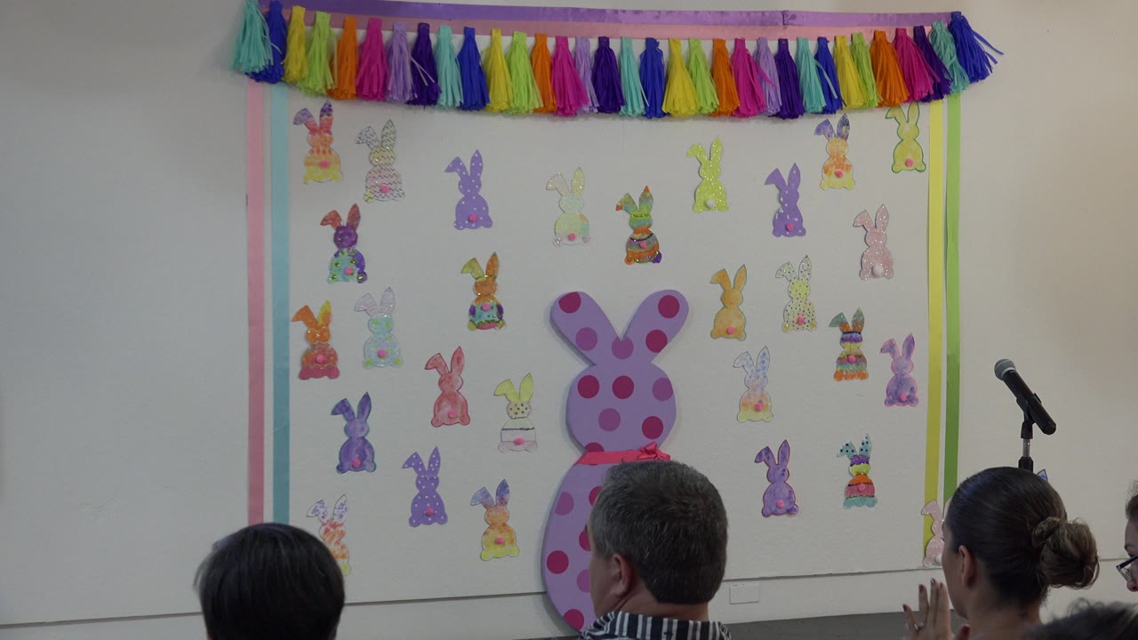 Audience members in a Cabo San Lucas classroom clap while facing a wall of paper bunny decorations and a large pink bunny cutout near a microphone stand.