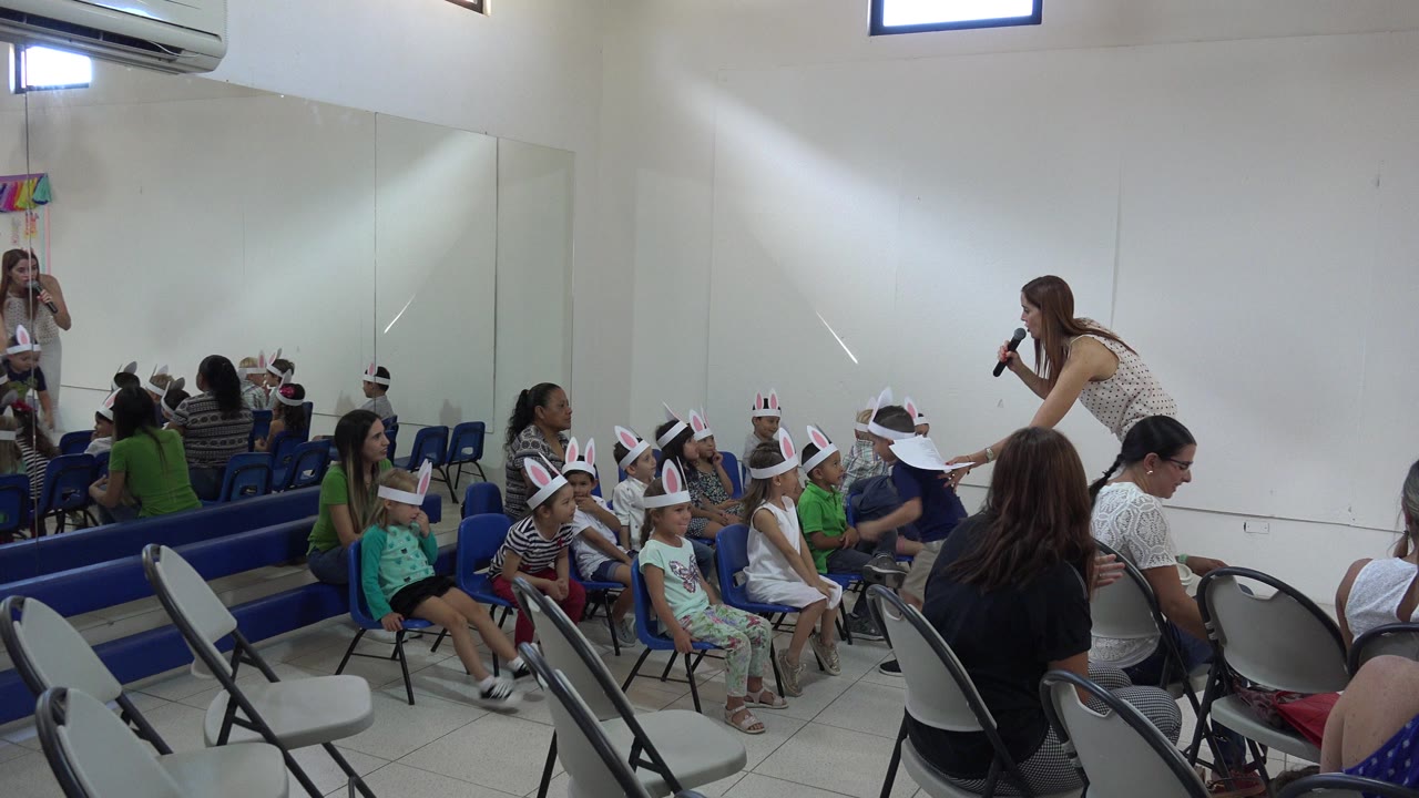 Woman with microphone leaning toward seated children in bunny ears.