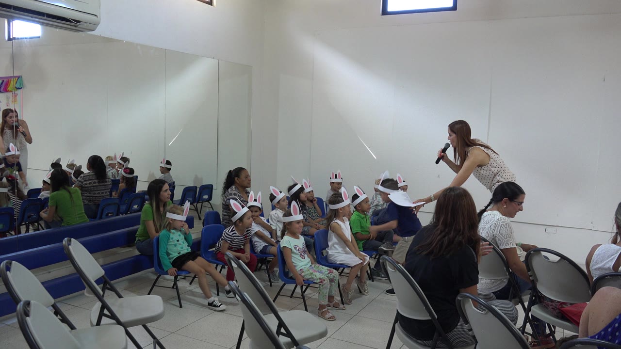 One second in, the woman remains bent toward the seated children as adults sit in folding chairs and the mirrored wall reflects part of the room in Cabo San Lucas.