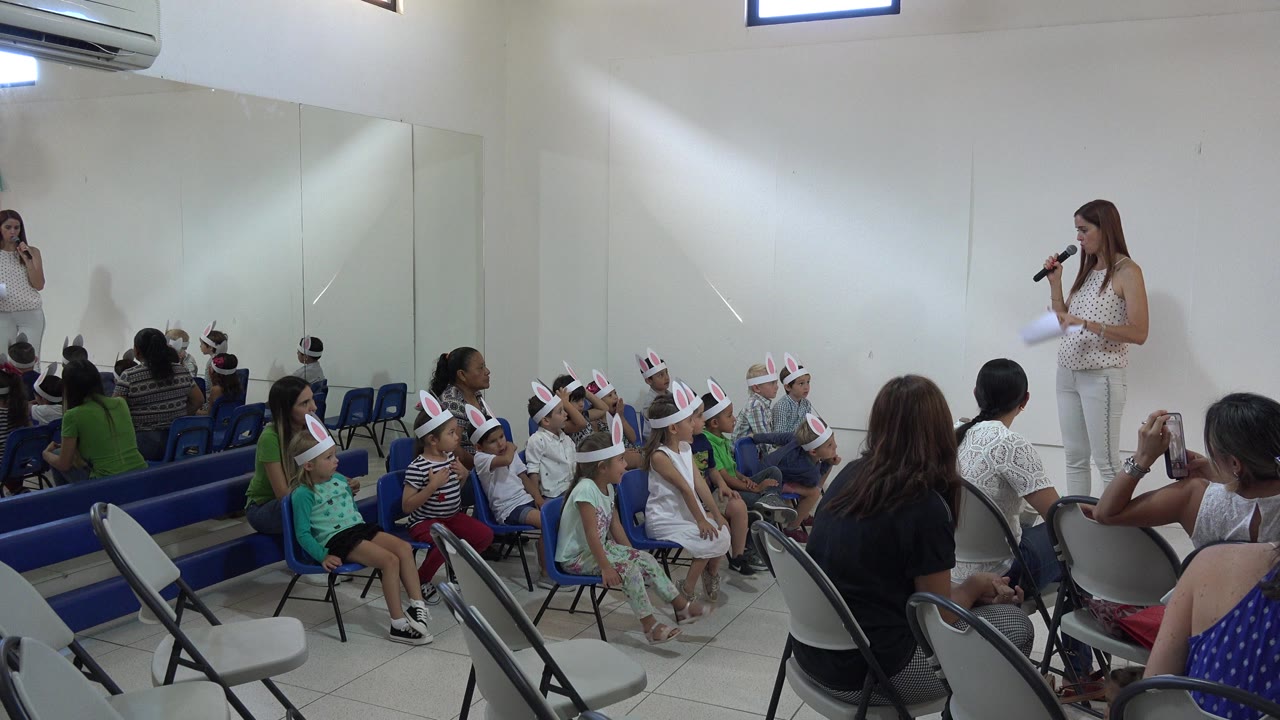 Children in rabbit-ear hats sit on blue chairs while a woman speaks at the front of the room.