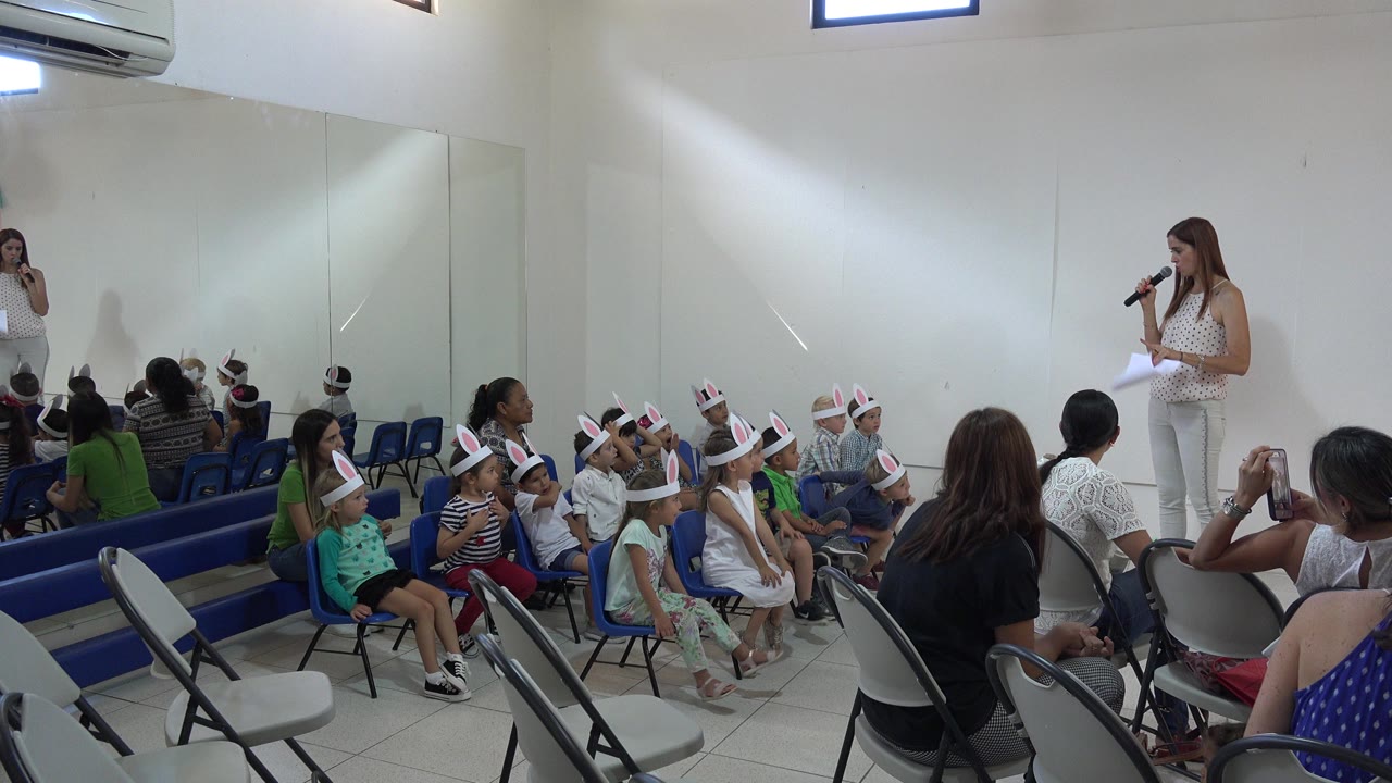 In Cabo San Lucas, Mexico, the presenter stands beside seated children in rabbit-ear hats while adults watch from folding chairs and blue seating rows.