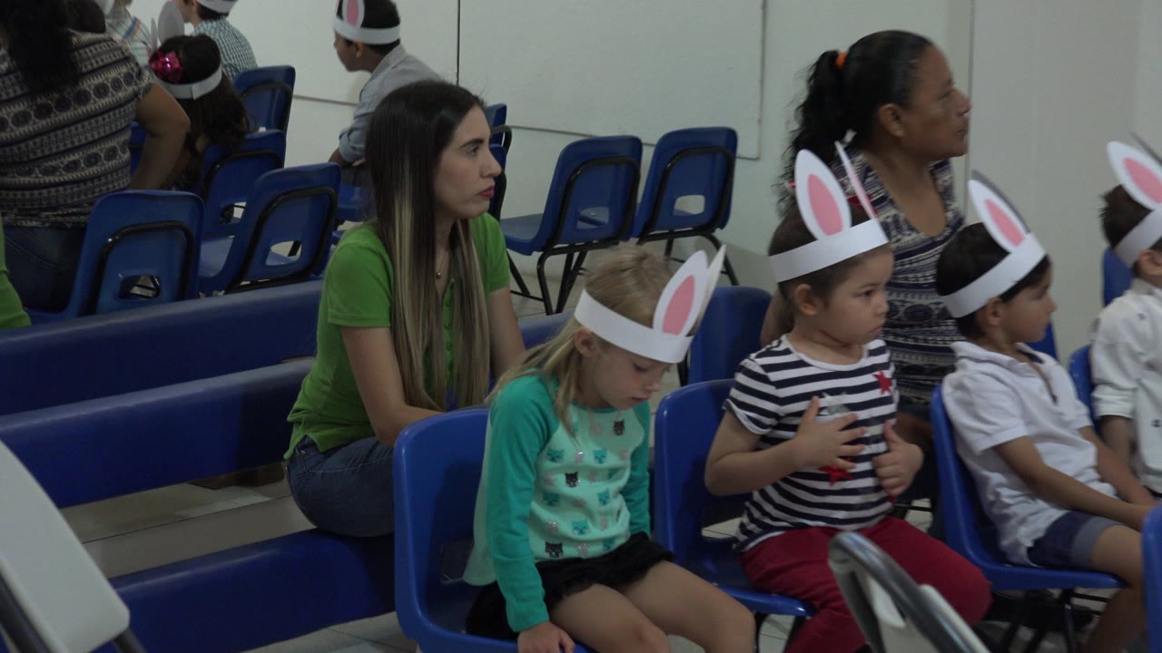 Children in bunny-ear headbands sit on blue classroom chairs.